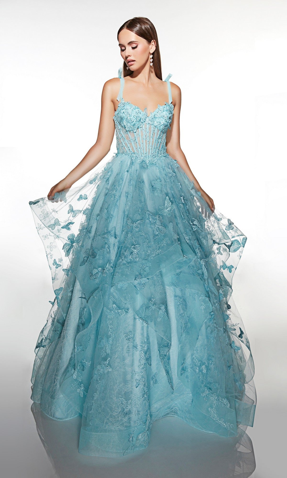 A woman poses against a white background in the Alyce 62199 long prom dress, a baby blue gown with floral and butterfly appliqué, a sheer corset bodice, spaghetti straps, and a tulle ballgown skirt.